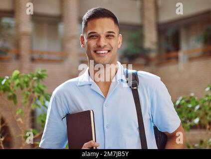 Portrait de face, homme et étudiant à l'université sur le campus prêt pour l'apprentissage. Éducation, université et heureux apprenant de sexe masculin du Brésil tenant livre pour Banque D'Images