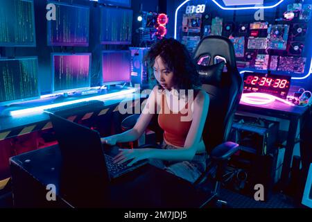 Hacker asiatique féminin dans un lieu de refuge avec un environnement sombre et plusieurs écrans et câbles, essayant de se casser dans les serveurs de données et d'infecter le système avec un virus. Banque D'Images