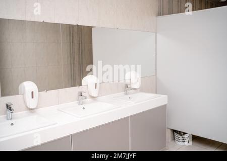 L'intérieur d'une toilette publique. Rangée de lavabos avec robinets en métal sur une dalle de marbre, distributeurs de savon liquide, miroir long sur un mur gris Banque D'Images