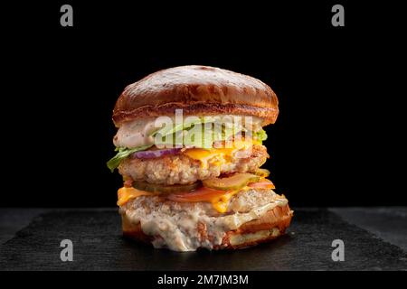 Hamburger avec deux côtelettes de poulet, fromage, cornichon, fromage, sauce et tomates, sur fond noir Banque D'Images