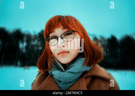 Portrait en gros plan d'une belle femme asiatique aux cheveux rouges vêtue d'un manteau beige qui marche dans un parc par une froide journée d'hiver. Une promenade en plein air le mois de janvier Banque D'Images