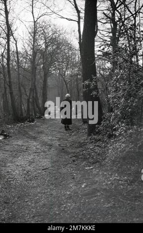 1955, historique, une jeune femme portant un long manteau en velours côtelé, un petit chapeau, des gants et des bottines marchant sur un chemin dans les bois, Farningham, Kent, Angleterre, Royaume-Uni. Banque D'Images