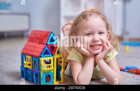 Jolie fille curly à côté d'une maison faite d'un constructeur magnétique à la maison sur le tapis dans la chambre des enfants. Banque D'Images