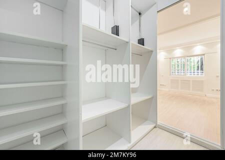 Une chambre vide aux murs peints en blanc avec accès à un dressing avec plusieurs étagères en marera blanc et des fenêtres en aluminium de style français Banque D'Images