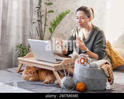 Une femme fait du crocheting tout en regardant un film sur un ordinateur portable. Apprenez à coudre à partir de leçons vidéo sur Internet. Maison confortable éclairée par la lumière du soleil. Banque D'Images