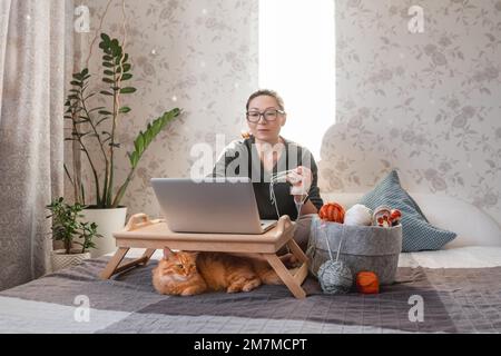 Une femme fait du crocheting tout en regardant un film sur un ordinateur portable. Apprenez à coudre à partir de leçons vidéo sur Internet. Maison confortable éclairée par la lumière du soleil. Banque D'Images
