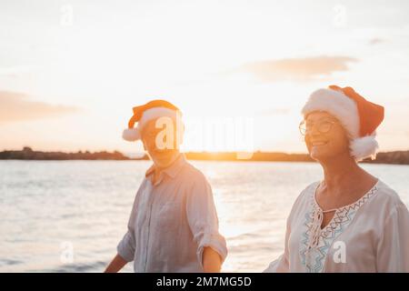 Vieux couple mignon de personnes matures appréciant et s'amusant ensemble à la plage portant des chapeaux de noël les jours de vacances. Marche sur la plage avec le coucher du soleil à l'arrière-plan en hiver. Banque D'Images