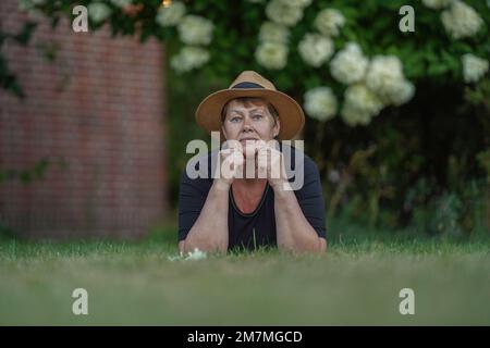 Une jeune grand-mère dans un chapeau de paille repose sur l'herbe dans le jardin avec son menton sur ses mains, vue du sol Banque D'Images