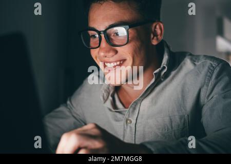 Gros plan et portrait d'un jeune adolescent étudiant et travaillant à la maison la nuit sur la table à l'aide d'un PC portable souriant et s'amusant à faire ses devoirs. Lumière de l'écran sur le visage. Banque D'Images