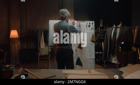 Homme mature tailleur dans un atelier de designer de luxe ou de tailleur. Il travaille avec le croquis du futur vêtement sur la table avec le tissu et les outils. Mannequin avec costume ajusté. Mode et artisanat. Banque D'Images