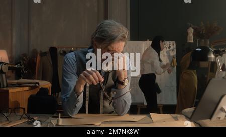 Un homme fatigué tailleur s'assoit à la table avec des outils de personnalisation, des motifs, une tablette et un ordinateur portable. Il prend des lunettes et pense. Femme tailleur sur la robe sur fond. Mode, style et artisanat. Banque D'Images