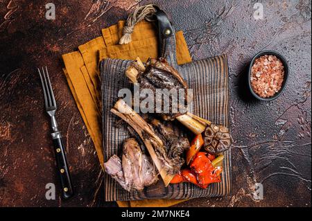 Branche d'agneau ou de jarret d'agneau cuit lentement et traditionnel avec des légumes sur une planche à découper. Arrière-plan sombre. Vue de dessus Banque D'Images