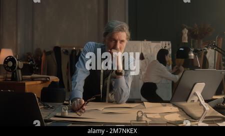 Un homme fatigué tailleur s'assoit à la table avec des outils de personnalisation, des motifs, une tablette et un ordinateur portable. Il prend des lunettes et pense. Femme tailleur sur la robe sur fond. Mode, style et artisanat. Banque D'Images