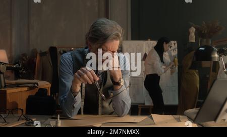 Un homme fatigué tailleur s'assoit à la table avec des outils de personnalisation, des motifs, une tablette et un ordinateur portable. Il prend des lunettes et pense. Femme tailleur sur la robe sur fond. Mode, style et artisanat. Banque D'Images