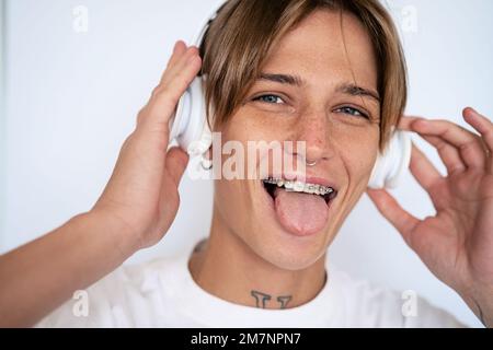 Femme blonde aux poils courts avec piercing facial et bretelles dentaires sur les dents écoutant de la musique avec des écouteurs et la langue. Banque D'Images