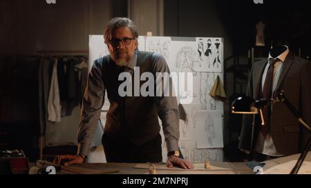 Homme mature tailleur travaille dans un atelier de designer de luxe ou en studio de couture, regarde la caméra. Table avec fils, tissu et outils. Combinaison et planche sur mesure avec croquis sur fond. Mode concept. Banque D'Images