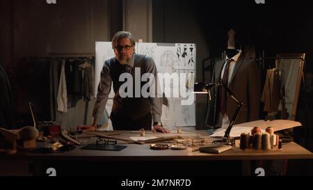 Homme mature tailleur travaille dans un atelier de designer de luxe ou en studio de couture, regarde la caméra. Table avec fils, tissu et outils. Combinaison et planche sur mesure avec croquis sur fond. Mode concept. Banque D'Images