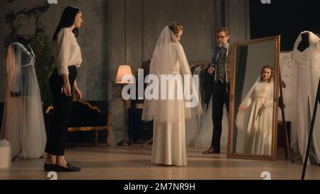 Photo cinématographique de femme tailleur mettant le voile sur la femme dans la robe de mariage devant le miroir. Mariée sur la tenue de mariage dans l'atelier de designer ou le salon de mariage. Concept de mode et de couturier. Banque D'Images