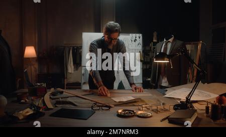 Homme mature tailleur dans un atelier de designer de luxe ou de tailleur. Il travaille avec le croquis du futur vêtement sur la table avec le tissu et les outils. Mannequin avec costume ajusté. Mode et artisanat. Banque D'Images