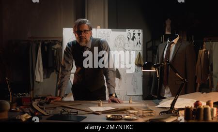 Homme mature tailleur travaille dans un atelier de designer de luxe ou en studio de couture, regarde la caméra. Table avec fils, tissu et outils. Combinaison et planche sur mesure avec croquis sur fond. Mode concept. Banque D'Images