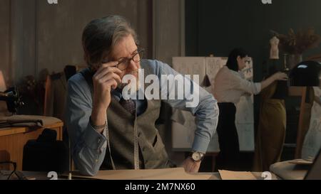 Un homme fatigué tailleur s'assoit à la table avec des outils de personnalisation, des motifs, une tablette et un ordinateur portable. Il prend des lunettes et pense. Femme tailleur sur la robe sur fond. Mode, style et artisanat. Banque D'Images