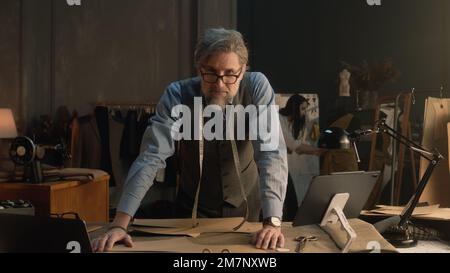 Un homme mature crée des vêtements pour la nouvelle collection de vêtements à l'aide d'une tablette et d'un ordinateur portable. Des lunettes sur mesure permettent de regarder l'appareil photo, de se trouver près de la table avec des outils de personnalisation. Concept de mode et d'artisanat. Banque D'Images