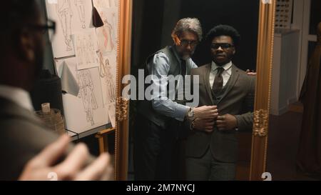 Homme afro-américain en costume dans un atelier de designer élégant ou dans une boutique de vêtements de luxe sombre. Tailleur met la veste sur l'homme devant le miroir. Costume de mariage ou de travail sur mesure. Mode concept. Banque D'Images