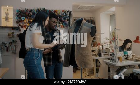 Un designer de vêtements afro-américain tient à portée de main une tablette, montre le croquis pour personnaliser et discute de la combinaison. Les couturiers assis et travaillant aux machines à coudre. Rack de threads en arrière-plan. Atelier atelier. Banque D'Images