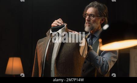 Homme mature tailleur travaille sur une combinaison élégante d'affaires ou de mariage. Mannequin avec chemise, cravate et veste sur mesure dans un atelier de designer de luxe élégant ou un atelier de tailleur sombre. Mode et artisanat. Banque D'Images