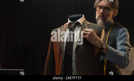 Homme mature tailleur travaille sur une combinaison élégante d'affaires ou de mariage. Mannequin avec chemise, cravate et veste sur mesure dans un atelier de designer de luxe élégant ou un atelier de tailleur sombre. Mode et artisanat. Banque D'Images