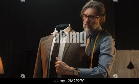 Homme mature tailleur travaille sur une combinaison élégante d'affaires ou de mariage. Mannequin avec chemise, cravate et veste sur mesure dans un atelier de designer de luxe élégant ou un atelier de tailleur sombre. Mode et artisanat. Banque D'Images