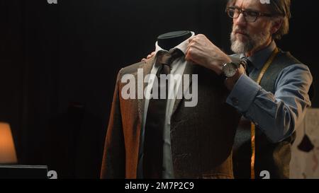 Homme mature tailleur travaille sur une combinaison élégante d'affaires ou de mariage. Mannequin avec chemise, cravate et veste sur mesure dans un atelier de designer de luxe élégant ou un atelier de tailleur sombre. Mode et artisanat. Banque D'Images