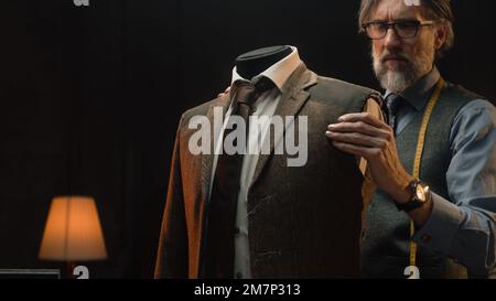 Homme mature tailleur travaille sur une combinaison élégante d'affaires ou de mariage. Mannequin avec chemise, cravate et veste sur mesure dans un atelier de designer de luxe élégant ou un atelier de tailleur sombre. Mode et artisanat. Banque D'Images