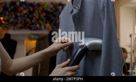 Gros plan d'une femme redonne une chemise ajustée avec défroisseur électrique à main dans un atelier d'atelier. Un tailleur pour homme rade les vêtements sur le fond. Concept de petite entreprise, industrie de la couture et mode. Banque D'Images