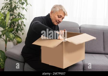 Belle femme senior tient une boîte en carton assise sur un canapé à la maison. Banque D'Images