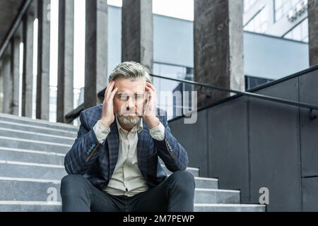 Homme d'affaires aux cheveux gris assis triste dans les escaliers à l'extérieur de l'immeuble de bureaux, patron déprimé en costume d'affaires contrarié et déçu par les résultats de réalisation. Banque D'Images