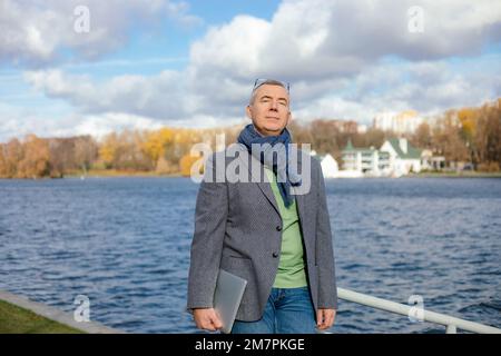 Vieux homme d'affaires dans des vêtements élégants appréciant avec les yeux fermés beauté naturelle. L'homme marche à l'extérieur près de l'étang d'eau. Banque D'Images