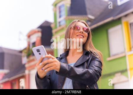 Jeune femme blonde en cuir veste et lunettes de soleil souriant avec le mobile, derrière la façade colorée Banque D'Images