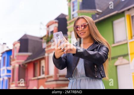 Jeune femme blonde en cuir veste et lunettes de soleil souriant avec le mobile, derrière la façade colorée Banque D'Images