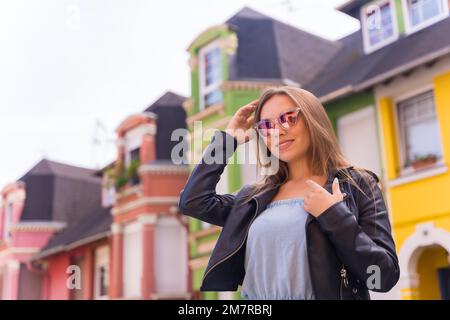 Posé de jeune femme blonde dans une veste en cuir et des lunettes de soleil souriantes, derrière une façade colorée Banque D'Images