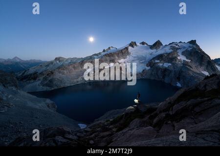 Homme debout sur une corniche au-dessus du lac alpin avec le phare allumé et la lune au loin Banque D'Images