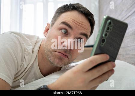 Homme surpris d'entendre la sonnerie de l'alarme de smartphone, se trouve dans le lit blanc. Réveillez-vous un gars choqué tard pour le travail le matin. Surprise du message sur smar Banque D'Images