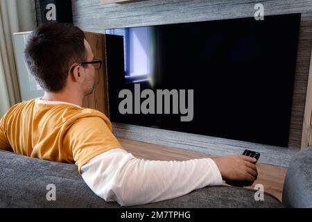 Homme regardant la télévision, allumant le téléviseur à écran plat plasma, pointant la télécommande vers un écran de télévision vide sur un mur blanc. Guy changer de chaîne à la maison Banque D'Images
