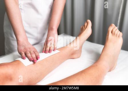 Esthéticienne cirant la jambe d'une femme appliquant une bande de matériel sur la cire chaude pour enlever les cheveux. Banque D'Images