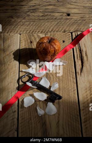 Une image de la vie avec un ruban rouge, des pétales de rose blancs, une vieille clé et une pomme pourrie posée sur de vieilles planches de bois. Banque D'Images