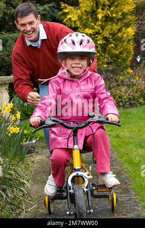 Un homme qui aide une jeune fille à faire son vélo jaune avec des stabilisateurs sur la voie dans un jardin Banque D'Images