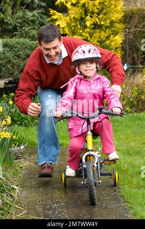 Un homme qui aide une jeune fille à faire son vélo jaune avec des stabilisateurs sur la voie dans un jardin Banque D'Images