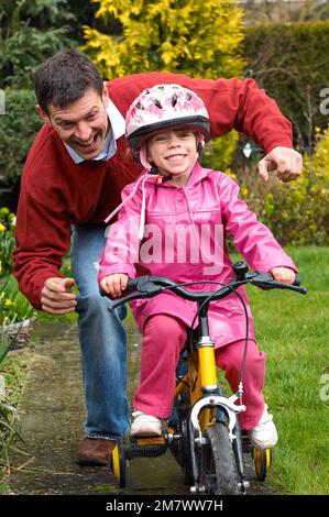 Un homme qui aide une jeune fille à faire son vélo jaune avec des stabilisateurs sur la voie dans un jardin Banque D'Images