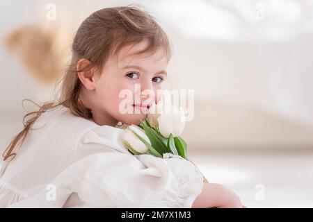 Portrait rapproché d'une petite fille dans une robe blanche, chemise de nuit. Le tout-petit embrasse un bouquet de tulipes blanches fraîches et délicates. Cadeau pour les vacances, le Banque D'Images