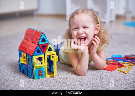 Portrait d'une fille en riant sur le tapis de la maison à côté d'une maison de jouet faite d'aimants. Banque D'Images